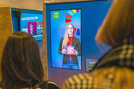 Am Eingang begrüßt Jugendaktivistin Greta das Publikum und informiert über die Nachhaltigkeitsziele der Vereinten Nationen Zwei Frauen stehen vor einer Videowand und sehen einem Film zu