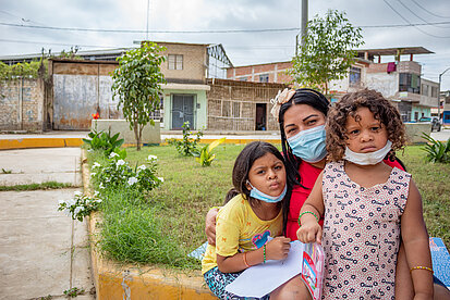 Eine junge Frau trägt einen Mundnasenschutz. Sie hält zwei junge Mädchen im Arm, beide schauen sehr ernst.