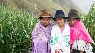 Drei Mädchen sitzen auf einer Bank. Sie tragen einen Rock, darüber ein Tuch und einen Hut. Sie grinsen in die Kamera. Hinter ihnen ist eine Berglandschaft zu sehen. 