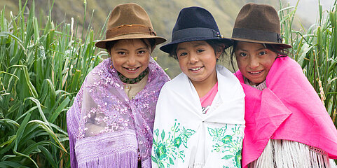 Drei Mädchen sitzen auf einer Bank. Sie tragen einen Rock, darüber ein Tuch und einen Hut. Sie grinsen in die Kamera. Hinter ihnen ist eine Berglandschaft zu sehen. 