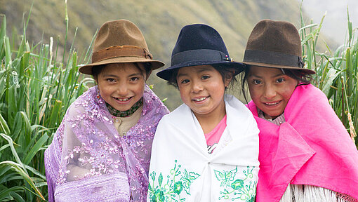 Evelyn Alexandra, 7 Jahre alt, lebt in Ecuador Drei Mädchen sitzen auf einer Bank. Sie tragen einen Rock, darüber ein Tuch und einen Hut. Sie grinsen in die Kamera. Hinter ihnen ist eine Berglandschaft zu sehen.