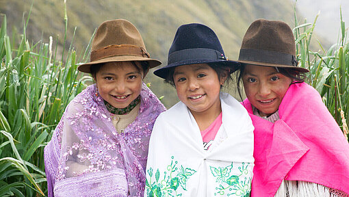Evelyn Alexandra, 7 Jahre alt, lebt in Ecuador Drei Mädchen sitzen auf einer Bank. Sie tragen einen Rock, darüber ein Tuch und einen Hut. Sie grinsen in die Kamera. Hinter ihnen ist eine Berglandschaft zu sehen.
