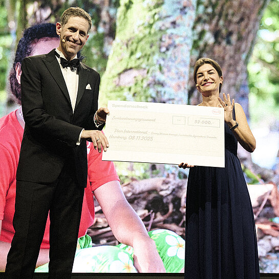 Eine Frau und ein Mann in eleganter Abendkleidung halten lächelnd einen Scheck über 92.000,- Euro in die Kamera.