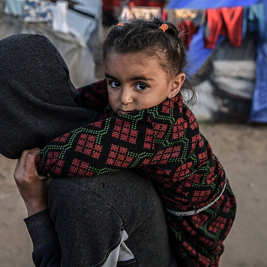 Kind in traditioneller Kleidung wird auf dem Rücken einer Person in Kapuzenjacke getragen, im Hintergrund Flüchtlingszelte in Gaza
