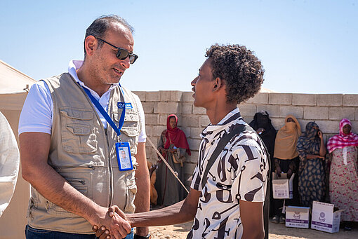 Mohamed Kamal (li.) beaufsichtigt Hilfslieferungen in Port Sudan Ein Mann mit Brille, Trackingjacke und Plan-Badge um den Hals schüttelt einem jungen Mann die Hand