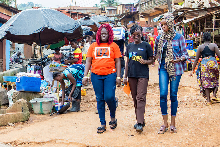 Drei junge Frauen laufen gemeinsam über eine Marktstraße in Freetown, Sierra Leone