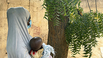 Eine junge Mutter mit langem Kopftuch hält ein Kind im Arm, sie ist seitlich von hinten fotografiert und nicht zu erkennen.