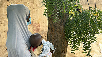 Eine junge Mutter mit langem Kopftuch hält ein Kind im Arm, sie ist seitlich von hinten fotografiert und nicht zu erkennen.