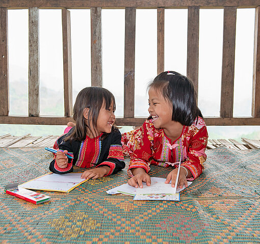 Zwei asiatisch gelesene Mädchen liegen auf dem Bauch und schreiben jeweils einen Brief. Sie lächeln sich an