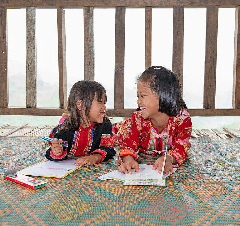 Zwei asiatisch gelesene Mädchen liegen auf dem Bauch und schreiben jeweils einen Brief. Sie lächeln sich an