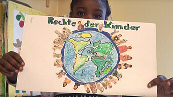 Ein Kind zeigt ein Blatt Papier in die Kamera, auf dem eine Weltkugel gemalt ist mit dem Text "Rechte der Kinder"