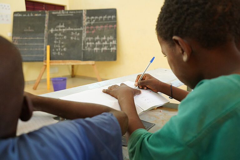 Zwei Jungen sitzen vor einer Tafel und schreiben in ihr Schulheft