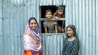 Zwei Mädchen in bunten Kleidern stehen vor einer Wellblechhütte, aus deren Fenster zwei kleine Kinder schauen