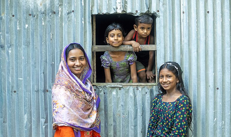 Zwei Mädchen in bunten Kleidern stehen vor einer Wellblechhütte, aus deren Fenster zwei kleine Kinder schauen