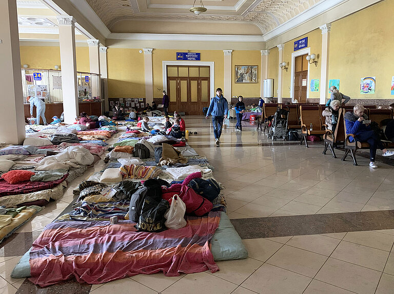 In einer Bahnhofshalle liegen Matratzen und Decken auf dem Boden, die als notdürftiger Schlafplatz für geflüchtete Menschen dienen.