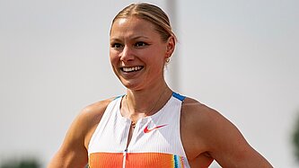 Porträt einer sportlich gekleideten Frau mit zurückgebundenen Haaren, die lächelt und ein ärmelloses Sportoberteil trägt. Der Hintergrund ist hell und unscharf.