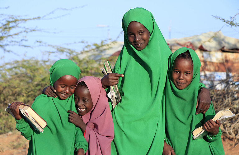 Vier Mädchen stehen eng beiinander, grinsen, und halten Schulbücher in der Hand. Drei tragen leuchtend grüne Gewänder, das vierte ein rosafarbenes. 