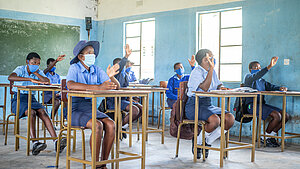 Schülerinnen und Schüler in Schuluniform und mit Masken sitzen in einem Klassenraum in Simbabwe und melden sich