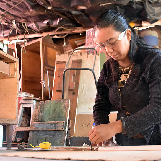 Frau mit Schutzbrille zeichnet in einer Werkstatt etwas auf einer Holzplatte