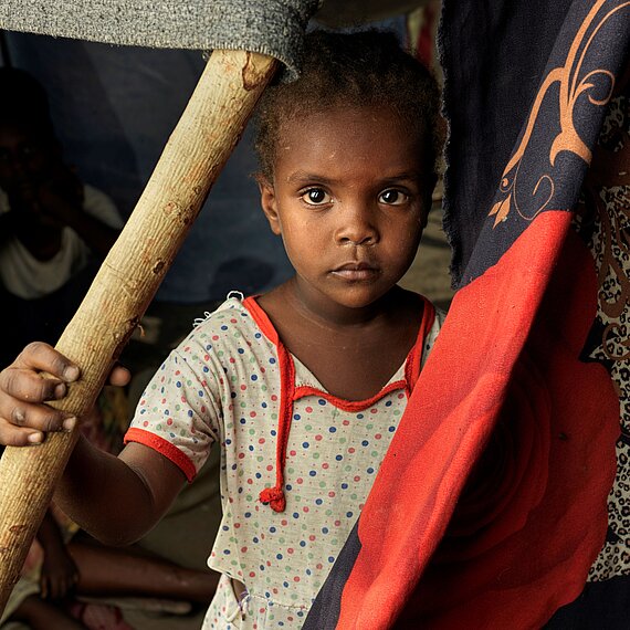 Ein junges Mädchen steht in einem provisorisch gebauten Zelt und hält sich an einer Stange fest. Sie blickt direkt in die Kamera.