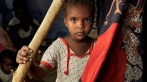 Ein junges Mädchen steht in einem provisorisch gebauten Zelt und hält sich an einer Stange fest. Sie blickt direkt in die Kamera.