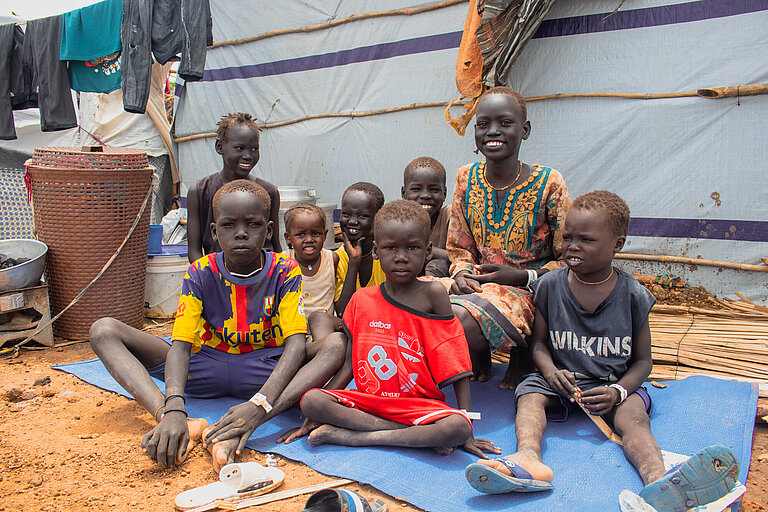 Acht Kinder unterschiedlichen Alters sitzen vor einem Shelter auf einer Decke. Einige Lachen, andere schauen ernst.