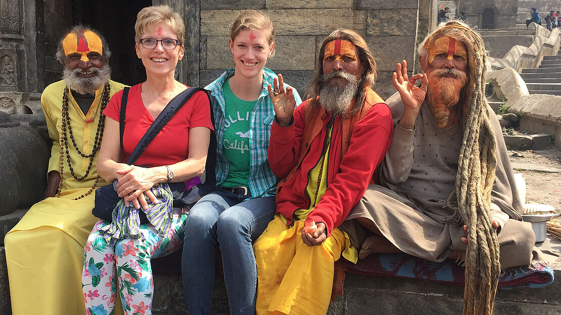 Henriette Kohlandt-Schönebeck und Annika Schönebeck sitzen neben traditionell gekleideten Männern mit langen Dreadlocks auf einer Steinmauer in Nepal