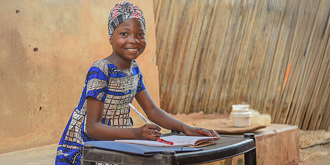 Ein Mädchen mit traditioneller, bunter Kleidung sitzt an einem kleinen Tisch. Vor ihr liegt ein Heft und sie hält einen Stift in der Hand. Sie lächelt in die Kamera.