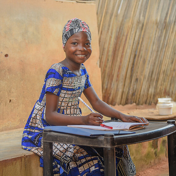 Ein Mädchen mit traditioneller, bunter Kleidung sitzt an einem kleinen Tisch. Vor ihr liegt ein Heft und sie hält einen Stift in der Hand. Sie lächelt in die Kamera.