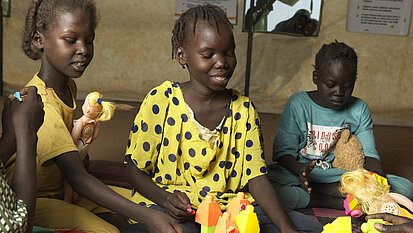 Drei Kinder sitzen auf einer Matte in einem Raum und spielen mit bunten Bauklötzen, einer Spielzeugpuppe und einem kleinen Spielzeugauto.