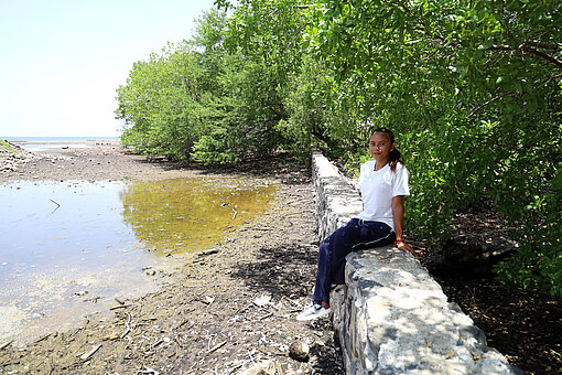 Junge Frau sitzt auf einer Steinmauer unter Bäumen neben dem Fluss