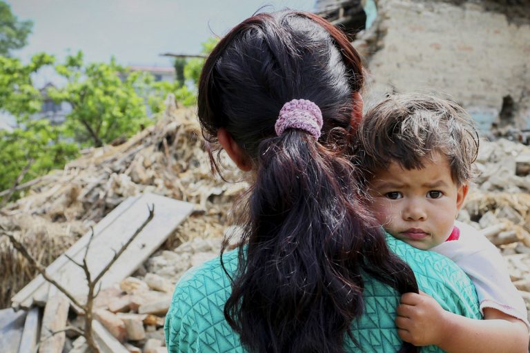 Frau mit Kind auf dem Arm steht vor Trümmern eines eingestürzten Hauses nach einem Erdbeben in Nepal. Das Kind blickt ernst in die Kamera.